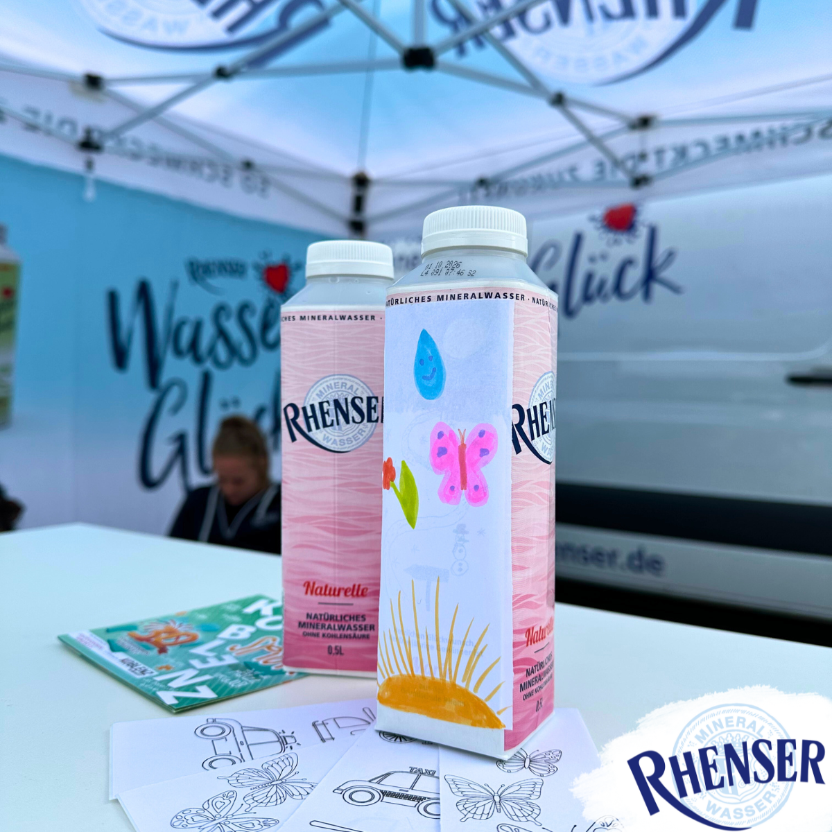 Zwei Rhenser Tetra-Top-Wasserflaschen, eine davon bunt bemalt, auf einem Tisch mit Ausmalbildern, im Hintergrund ein Promotion-Stand.