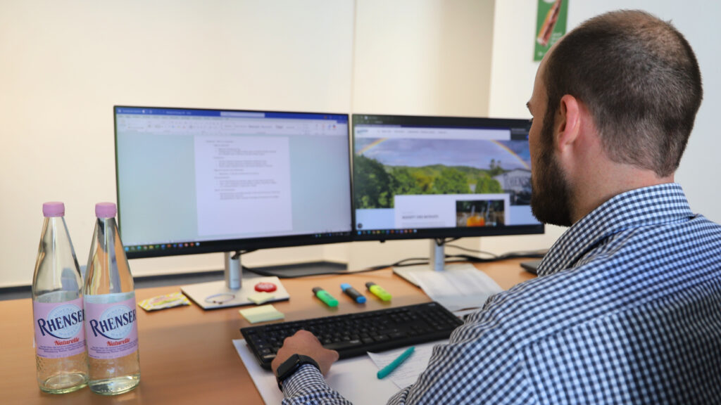 Mann arbeitet am Schreibtisch mit zwei Monitoren; auf dem Tisch stehen zwei Flaschen Rhenser Mineralwasser.