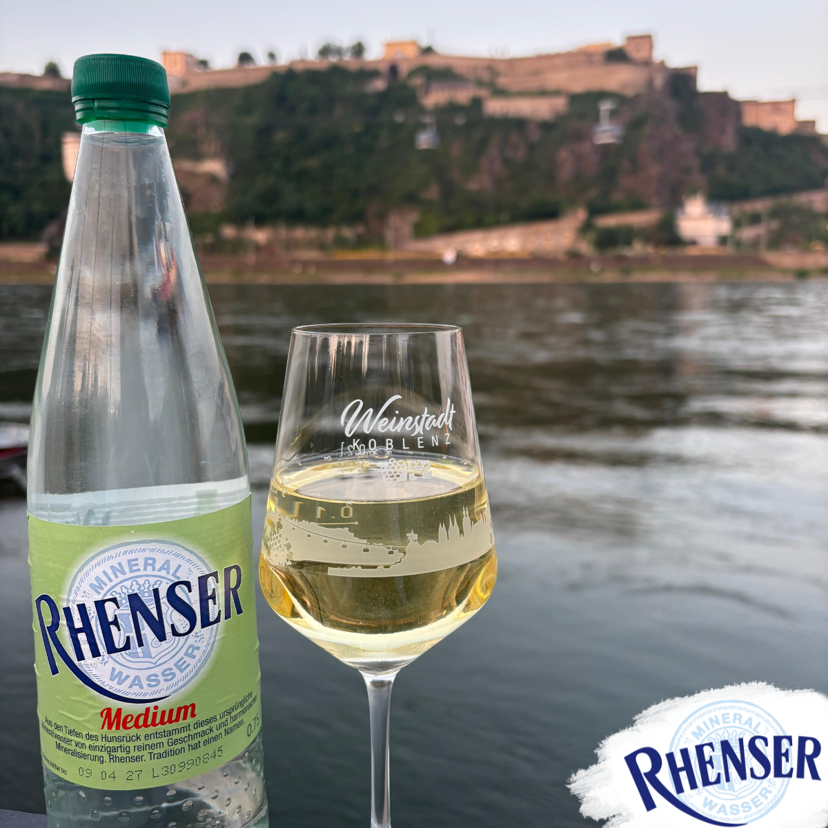 Glasflasche Rhenser Mineralwasser Medium neben Weinglas mit Weißwein und Aufdruck 'Weinstadt Koblenz' vor Rhein und Festung Ehrenbreitstein im Hintergrund.