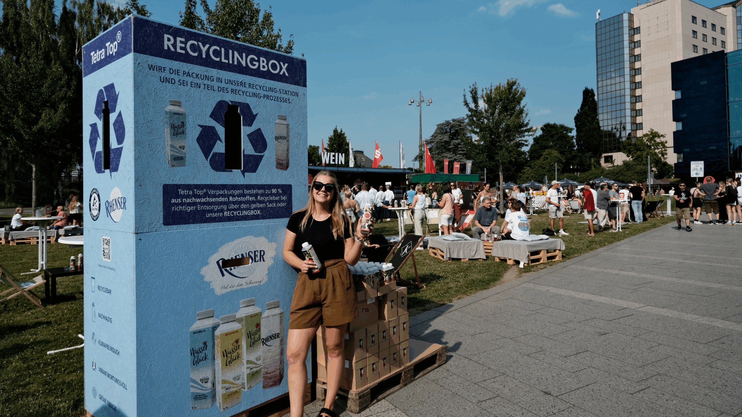 Rhenser Mitarbeiterin steht neben blauer Recyclingbox für Tetra-Top-Verpackungen auf einer grünen Wiese mit vielen Menschen im Hintergrund an einem sonnigen Tag an Rhein-Mosel-Halle Koblenz.