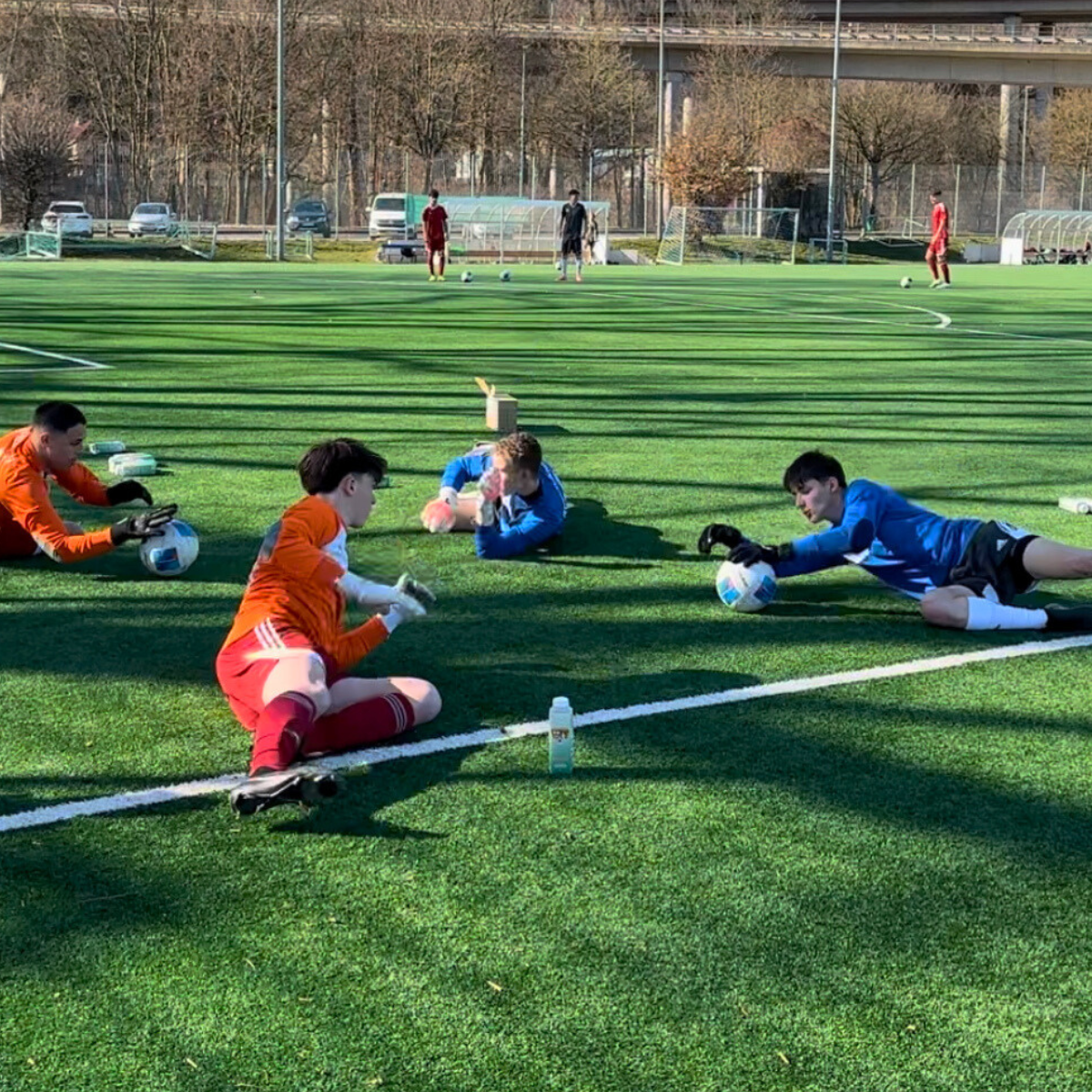 Vier Fußballtorhüter in Trainingskleidung liegen auf dem Rasen und halten jeweils einen Ball.