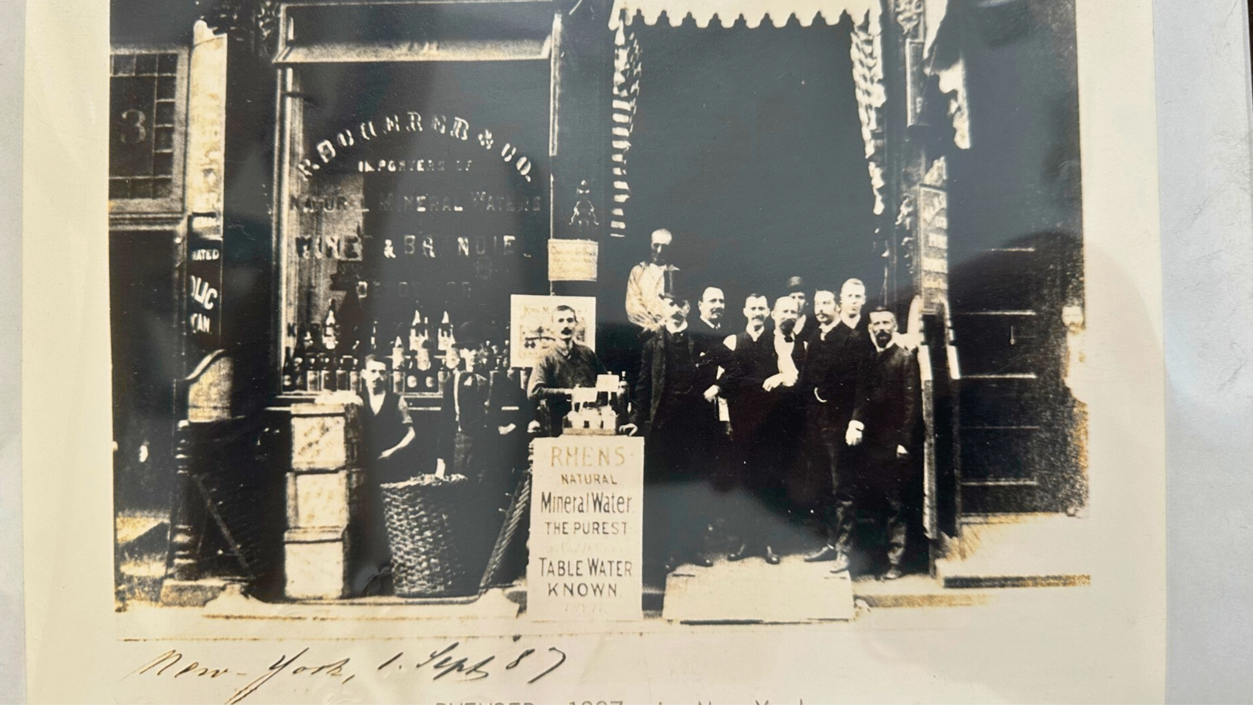 Schwarz-weiße-Foto einer Gruppe von Männern vor einem Geschäft in New York im Jahr 1887, mit einem Schild für „Rhens Natural Mineral Water“.