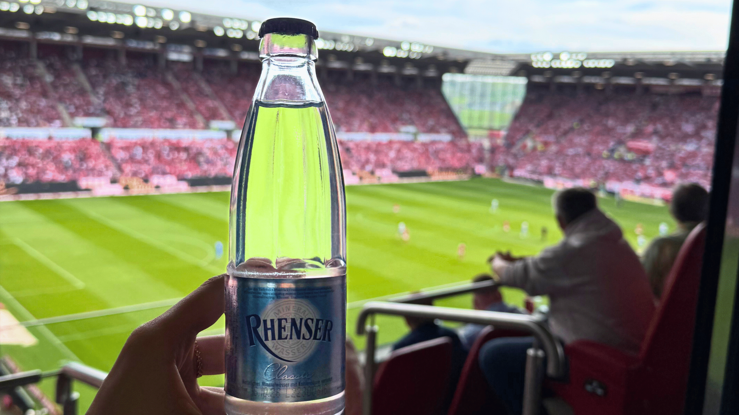Hand hält eine Flasche Rhenser Mineralwasser im Stadion mit Blick auf ein Fußballspiel.
