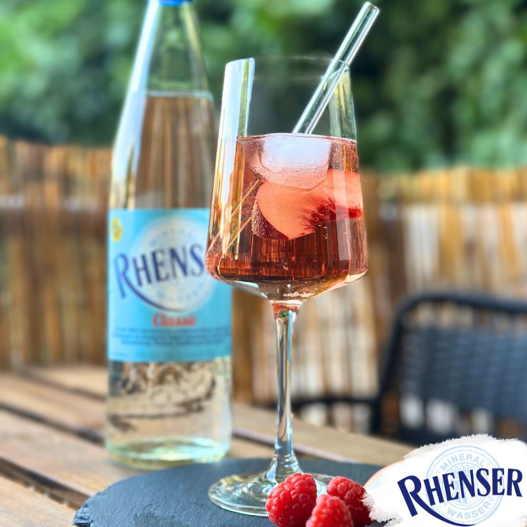 Ein Weinglas mit Pfirsich-Himbeer-Spritz mit Eiswürfeln, daneben eine Flasche Rhenser Mineralwasser Classic und drei Himbeeren auf schwarzer Platte.