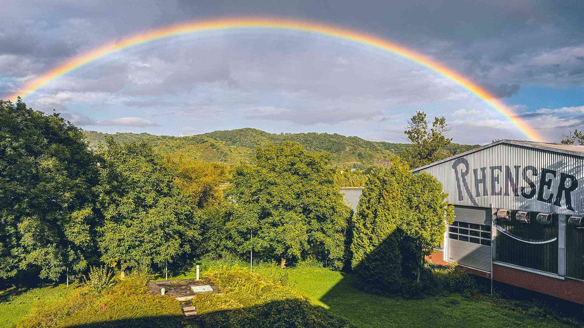 Regenbogen über grünen Hügeln und Bäumen neben dem Rhenser-Gebäude am Rhein.