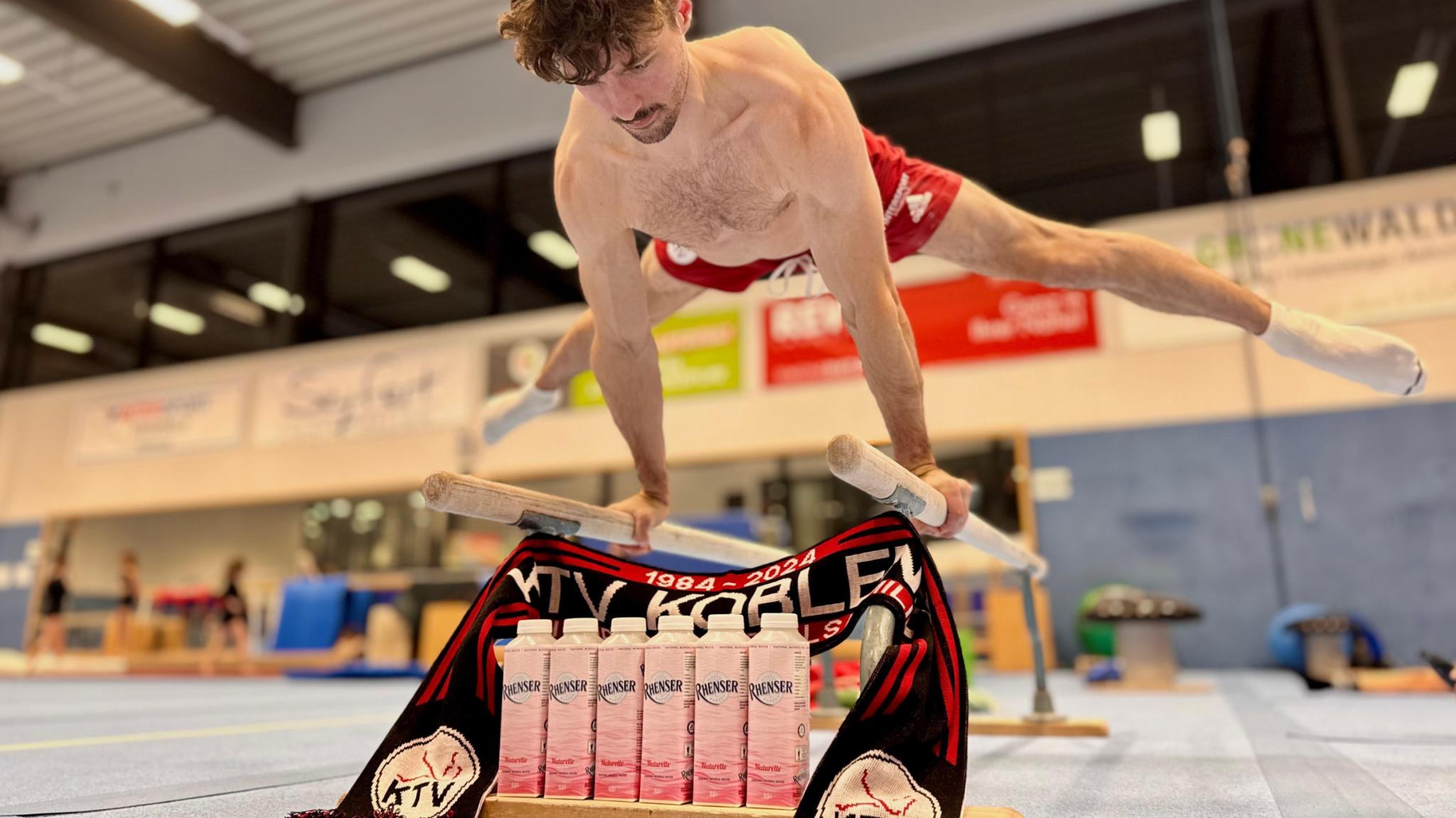 Ein Turner der KTV Koblenz zeigt eine Kraftübung am Barren; davor stehen mehrere Rhenser-Getränkekartons zusammen mit einem KTV-Schal in der Halle.