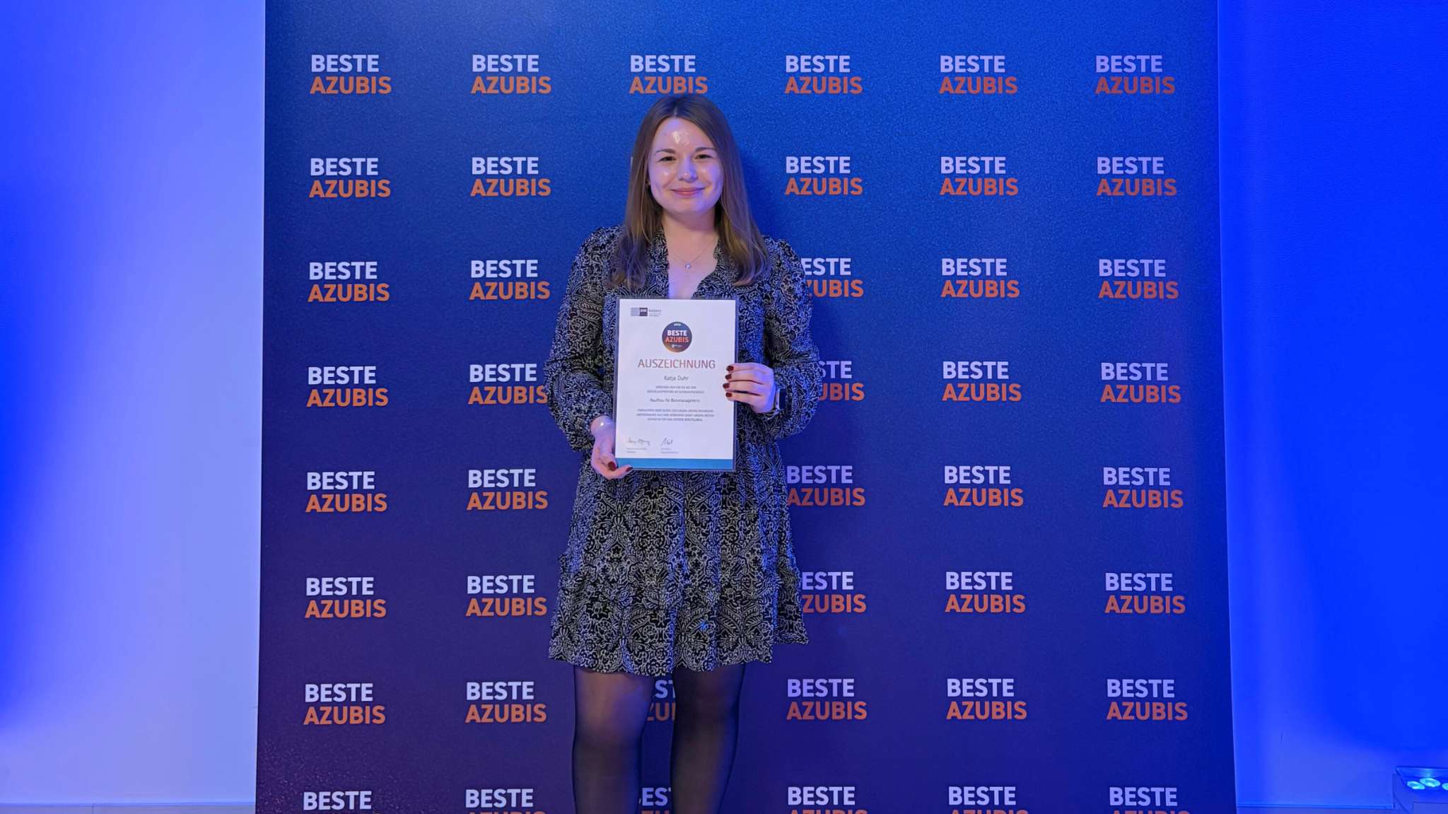 Katja Duhr hält IHK-Auszeichnung vor einer blau beleuchteten Wand mit dem Schriftzug ‘Beste Azubis’.