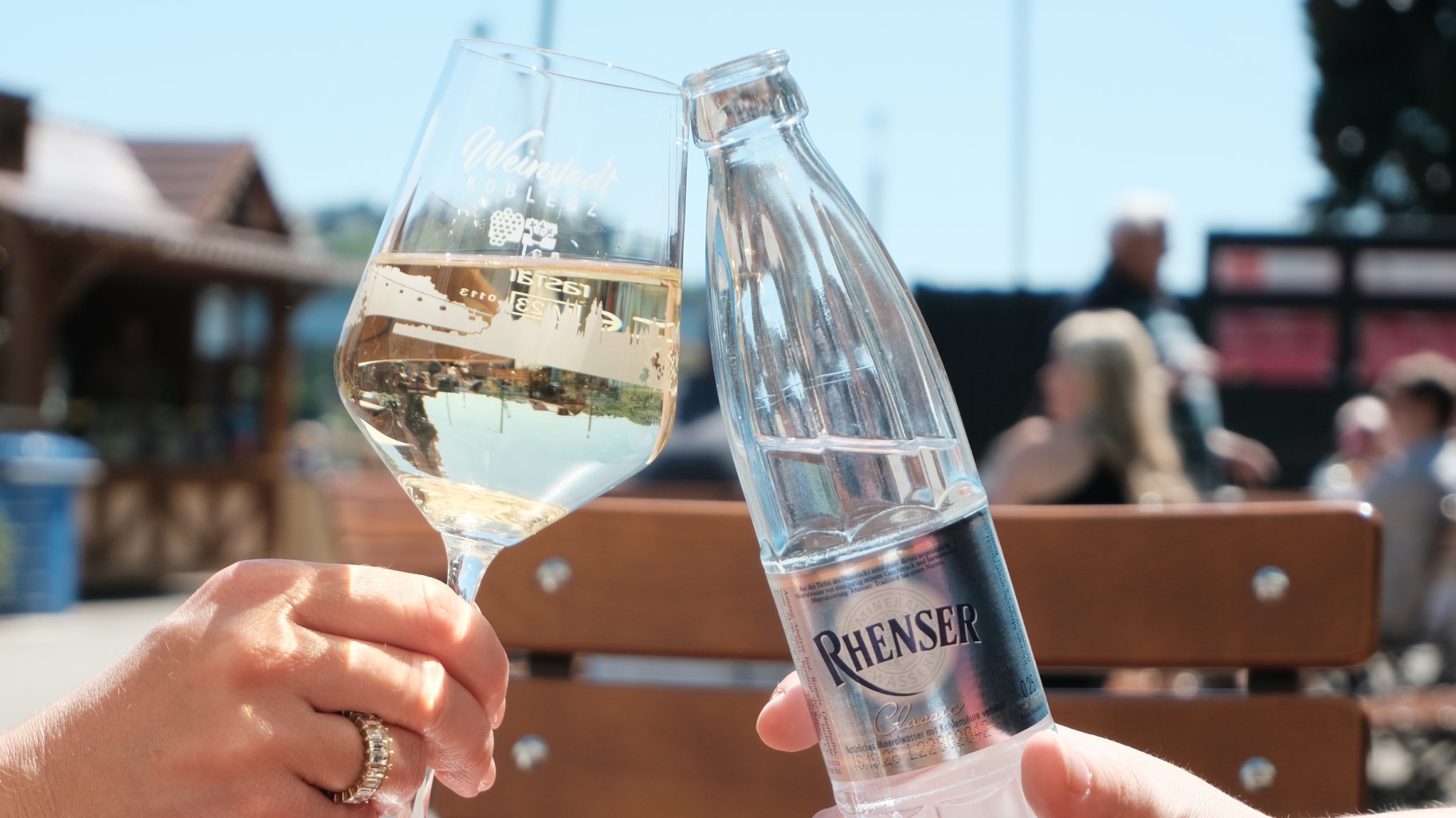 Zwei Personen stoßen mit einem Weinglas und einer Rhenser-Mineralwasserflasche im Freien an.