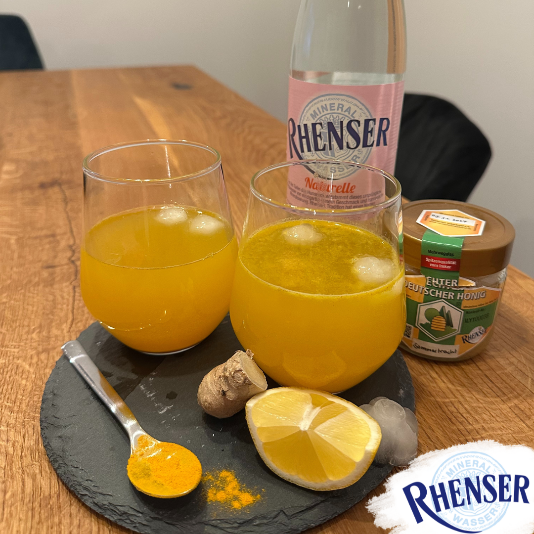 Zwei Gläser mit einem Orangenen Getränk. Im Hintergrund eine Rhenser Mineralwasser Flasche und im Vordergrund liegt eine Zitrone und Ingwer auf einer schwarzen Schieferplatte.