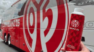 Eine Hand hält eine rote Mainz-05-Flasche vor einem Mannschaftsbus mit großem Vereinslogo.