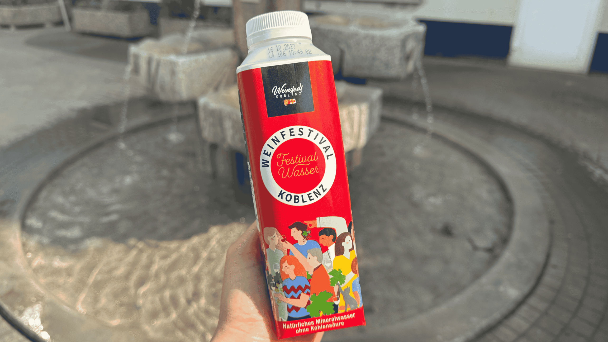 Eine Tetra-Top-Flasche im Festivalwasser-Design des Weinfestivals Koblenz wird vor einem Brunnen gehalten.