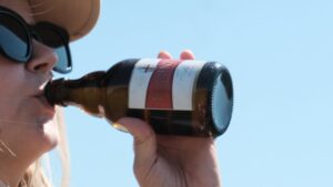 Eine Frau mit Sonnenbrille trinkt aus einer Flasche Koblenzer Bier vor blauem Himmel.