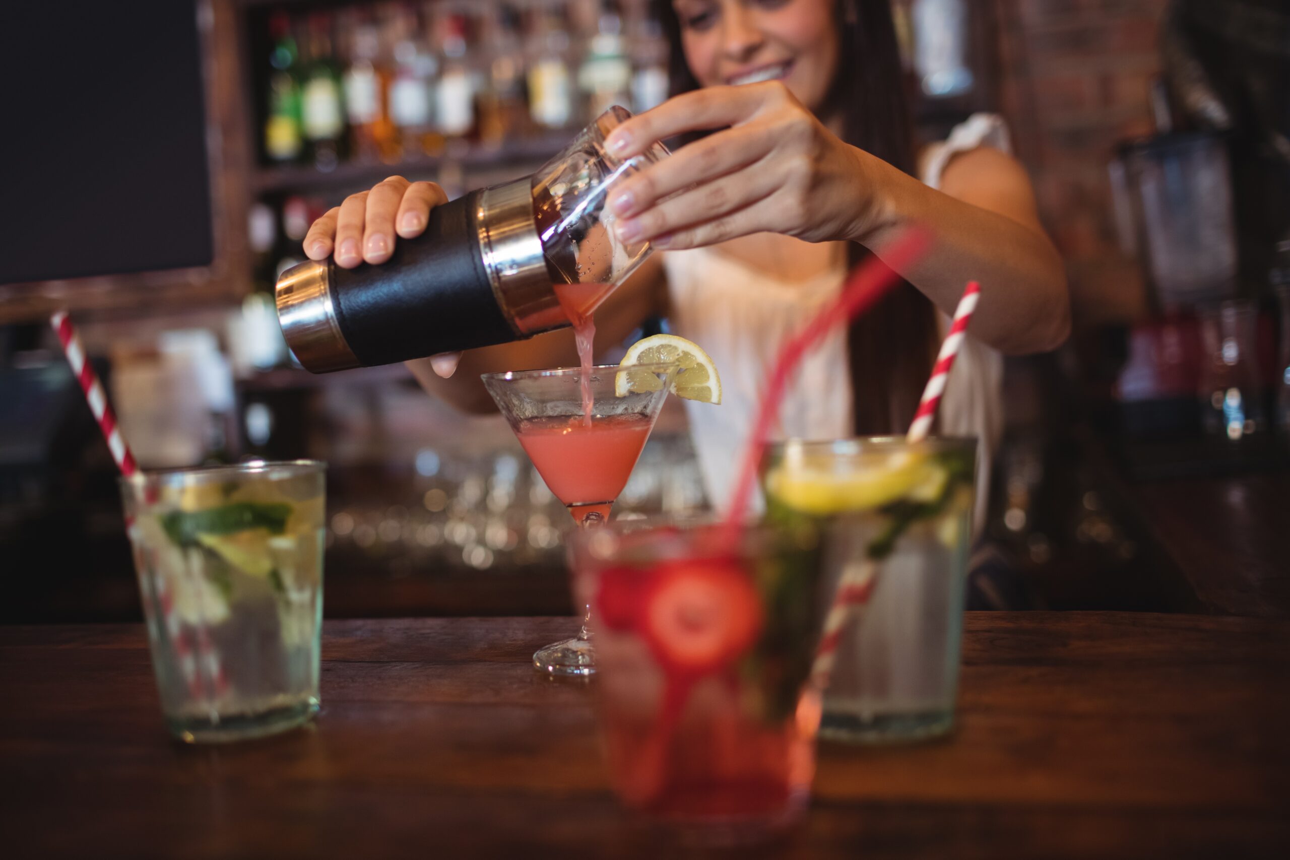Eine Frau gießt ein Cocktail in ein Cocktailglas an einer Bar. Daneben stehen noch weitere bunte Cocktails.
