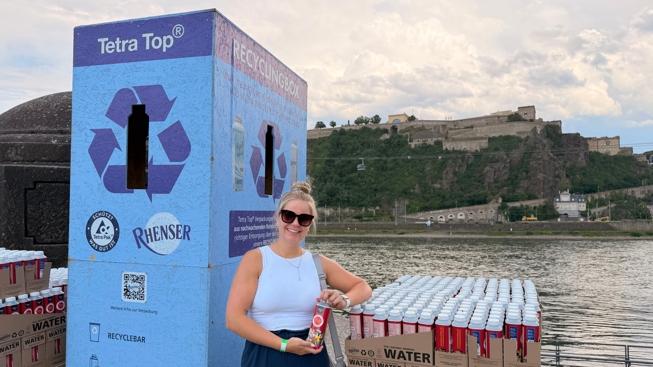 Person mit Rhenser Festivalwasser vor einer Rhenser Recyclingbox am Rheinufer in Koblenz, im Hintergrund die Festung Ehrenbreitstein.