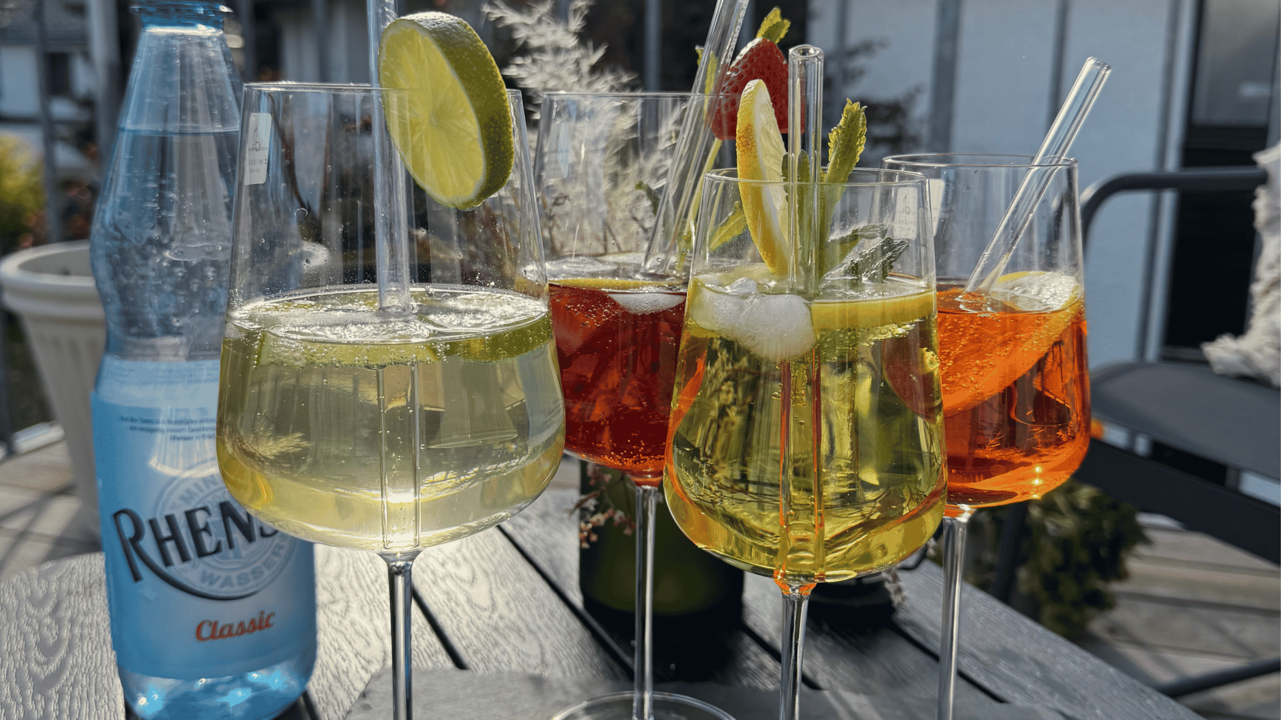 Rhenser Flasche Classic und 4 Weingläser mit verschiedenen bunten Spritz getränken, dekoriert auf einer Schieferplatte in einem grünen garten.