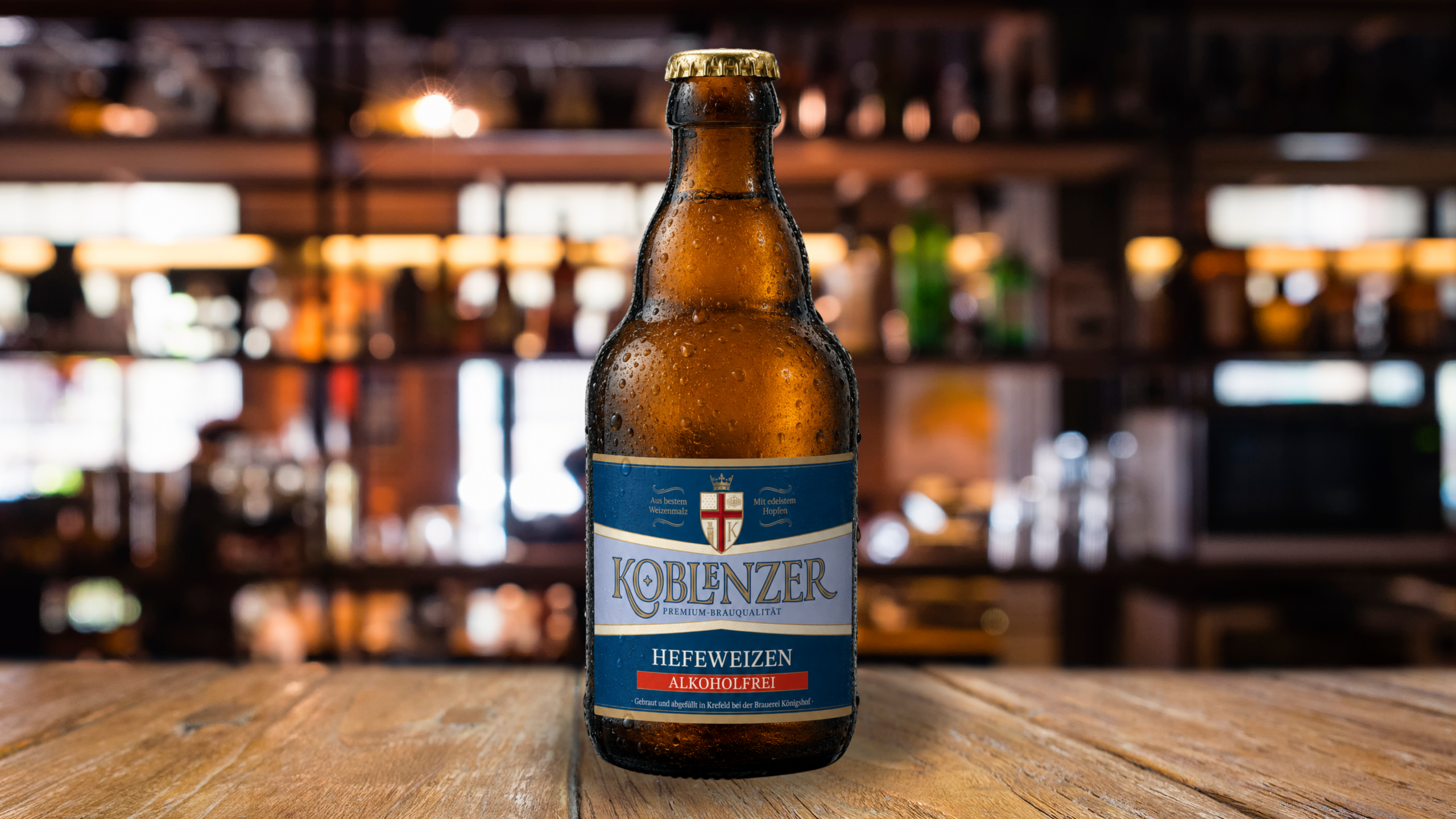 Flasche Koblenzer Hefeweizen alkoholfrei auf einem Holztisch vor unscharfem Barhintergrund.